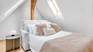Een moderne slaapkamer met een schuine kap, dakraam, een bed met veel kussens en een nachtkastje met een lamp.