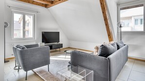 Een modern loft appartement met zichtbare houten balken, een grijze zithoek, een flatscreen televisie en een groot raam met uitzicht op de stad.