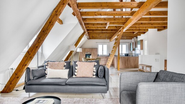 Een modern loft appartement met zichtbare houten balken, een grijze bank, een kleine ronde tafel en een keuken op de achtergrond.