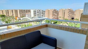 Un balcon doté d’un canapé en osier et d’une balustrade blanche, offrant une vue sur un paysage urbain avec de hauts immeubles et de la verdure.