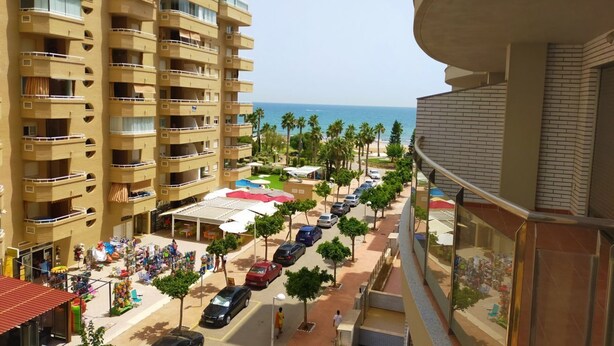 A multi-story building with balconies overlooking a street lined with parked cars and palm trees.