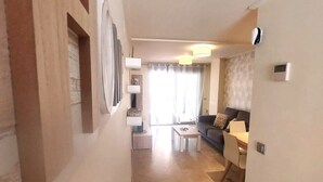A living room with a sofa, coffee table, and a dining area with chairs. There is a window with curtains, a lamp, and a wall-mounted air conditioner.
