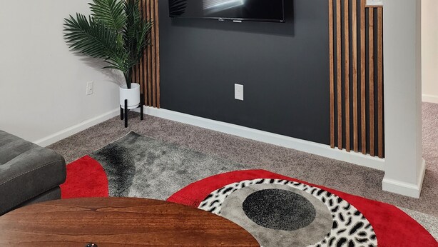 Una sala de estar moderna con un televisor de pantalla plana montado en una pared oscura, paneles de madera, un ventilador de techo, una planta en maceta y un tapete con estampado rojo y gris.