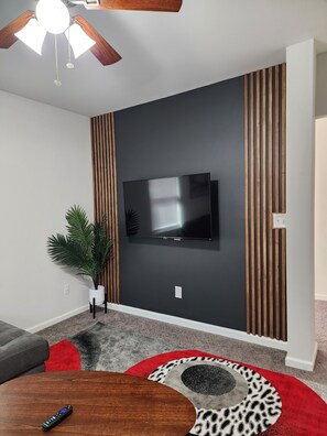 A modern living room with a flat-screen TV mounted on a dark wall, wooden paneling, a ceiling fan, a potted plant, and a red and grey patterned rug. - Quiet spacious 1BR Centrally Located w Parking/Washer/Dryer onsite. Hi Sp Wifi! (Hanahan)