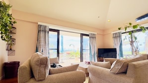 Ein Wohnzimmer mit einer Sofagarnitur, einem Couchtisch und einem Fernseher. Große Fenster mit Vorhängen, eine Pflanze und ein Blick ins Freie.