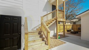 Una propiedad residencial con una escalera de madera que conduce a un porche cubierto, una puerta principal negra y un patio trasero cercado con mobiliario exterior.