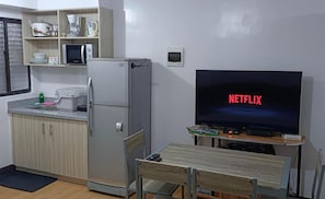 A compact kitchen with a refrigerator, microwave, and coffee maker. A flat-screen TV displays "NETFLIX" and is placed on a wooden table. - 2-Bedroom Condo w/ Unli Wifi, Netflix, Prime Video, Apple TV, Game Console, Pool (Imus)