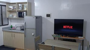 A compact kitchen with a refrigerator, microwave, and coffee maker. A flat-screen TV displays "NETFLIX" and is placed on a wooden table. - 2-Bedroom Condo w/ Unli Wifi, Netflix, Prime Video, Apple TV, Game Console, Pool (Imus)