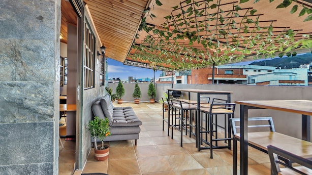 Een terras op het dak met een stenen muur, een grijze bank, houten tafels en plantenbakken.
