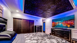 A bedroom with a starry ceiling, a large bed, a dresser, and a TV displaying a cityscape.