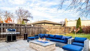 An outdoor patio with a stone fire pit, blue cushioned seating, and a grill.