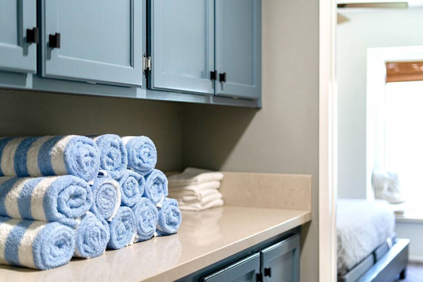 A neatly arranged stack of blue and white striped towels on a countertop, with a bed and a window in the background.