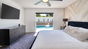 A bedroom with a bed, a ceiling fan, a TV, a nightstand, and a view of an outdoor swimming pool.
