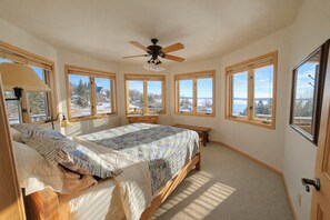 A bedroom with a bed, a desk, a chair, a ceiling fan, and large windows with a view.