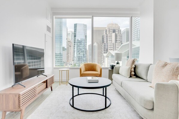 Relax in a spacious living room with a large window offering a view of high-rise buildings, a comfortable sofa, and a television on a stand.
