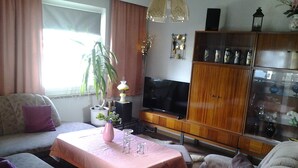 Un salon comprenant un canapé, une table recouverte d’une nappe rose, un téléviseur et un meuble avec des portes en verre.