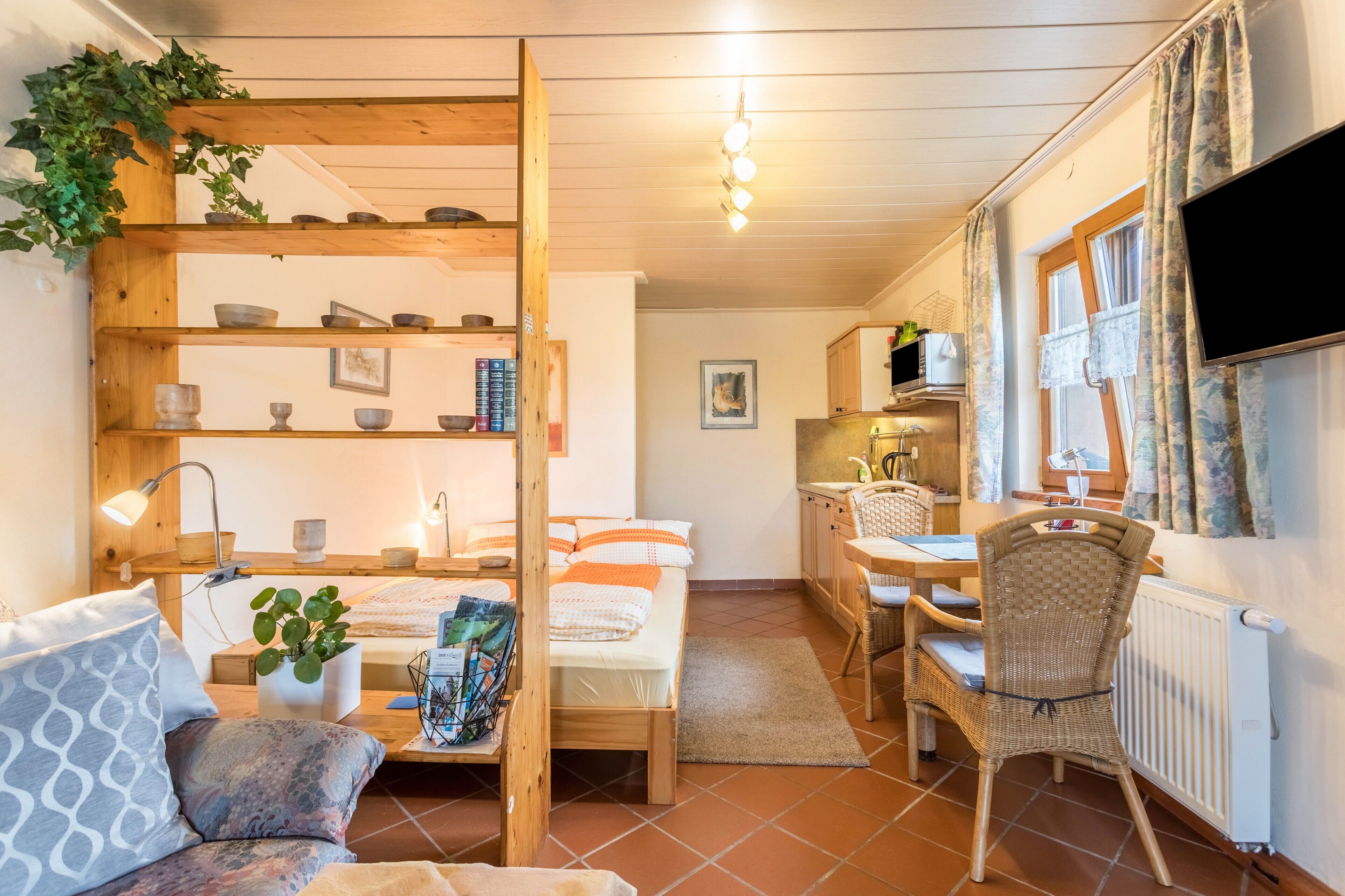 A cozy room with a bed, a desk, a chair, a shelf with bowls, and a TV.