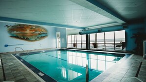 An indoor swimming pool with a mural on the wall, a view of the ocean through large windows, and a tiled floor. - BEACH LEVEL CONDO L08-2BR/2BA-Private Beach-Indoor Pool. (Geneva-on-the-lake)