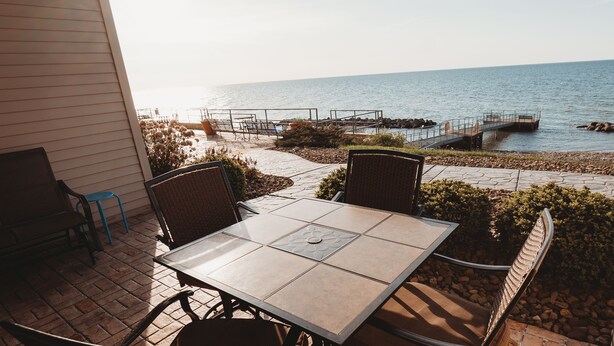 A patio with a table and chairs overlooking a rocky shoreline and a pier. - BEACH LEVEL CONDO L08-2BR/2BA-Private Beach-Indoor Pool. (Geneva-on-the-lake)