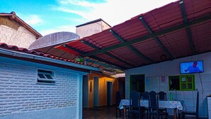 Un espace extérieur abrité, aménagé pour prendre les repas, avec un sol carrelé, une table et des chaises, et un téléviseur fixé au mur.