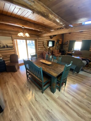 A log cabin interior with a wooden dining table, chairs, a sofa, and a television.
