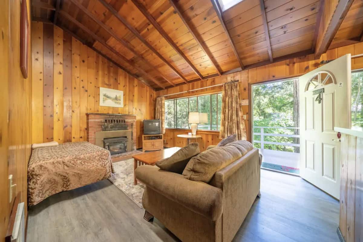 A cozy cabin interior with wooden walls and a fireplace. There is a sofa and a small table with a lamp.