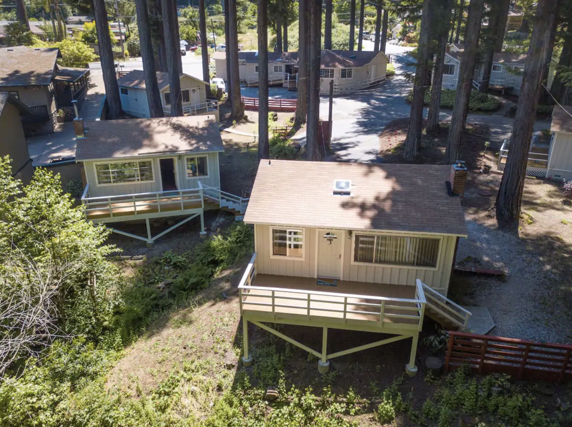 Deux petites cabanes avec vérandas attenantes, entourées de grands arbres et bordées d’une allée de gravier.
