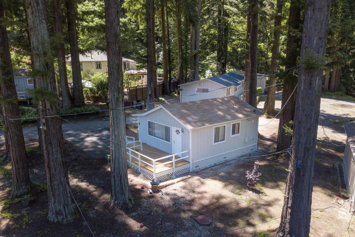 A small, single-story cabin with a porch, surrounded by tall trees in a wooded area.