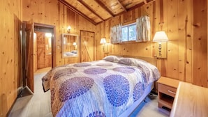 A wooden cabin bedroom with a large bed, a wooden dresser, and a window with curtains. - Masood's Lodge (Boulder Creek)