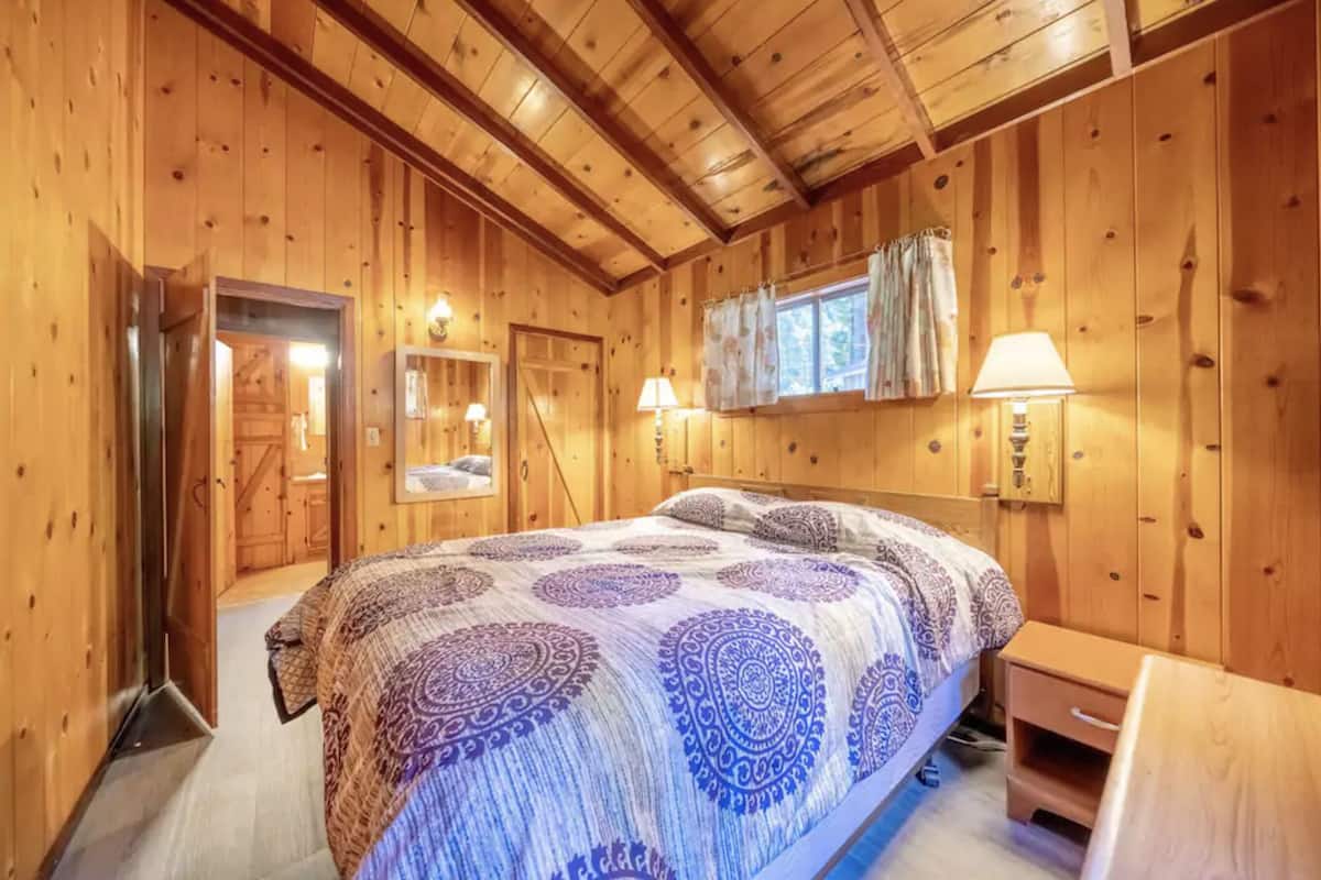 A wooden cabin bedroom with a large bed, a wooden dresser, and a window with curtains.