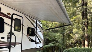A white RV with a large awning, parked in a forested area with a gravel path and a bench nearby. - Masood's Lodge (Boulder Creek)