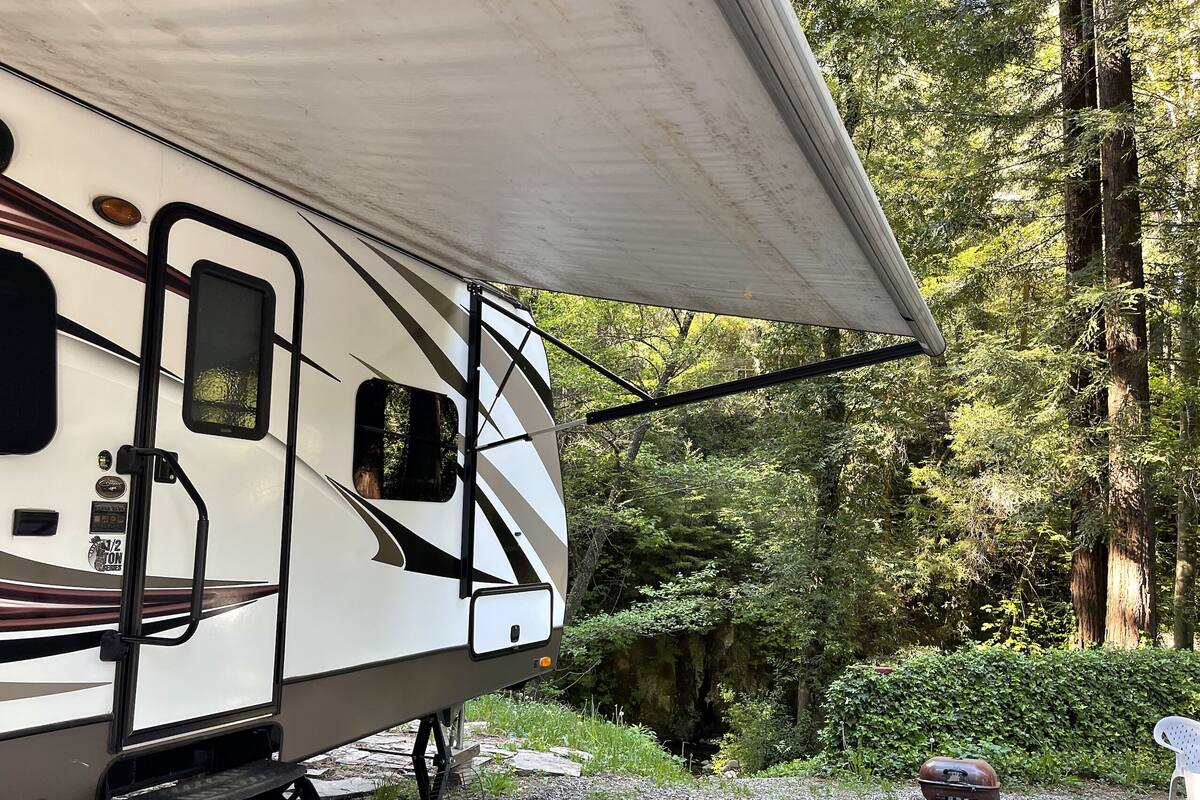 A white RV with a large awning, parked in a forested area with a gravel path and a bench nearby.