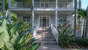 A two-story building with a front porch, a staircase leading to the entrance, and a well-maintained garden with tropical plants. - Luxury Harbor Home – Pool, Hot Tub, Basketball Court & Tiki Lounge Near Gulf Beaches (Largo)