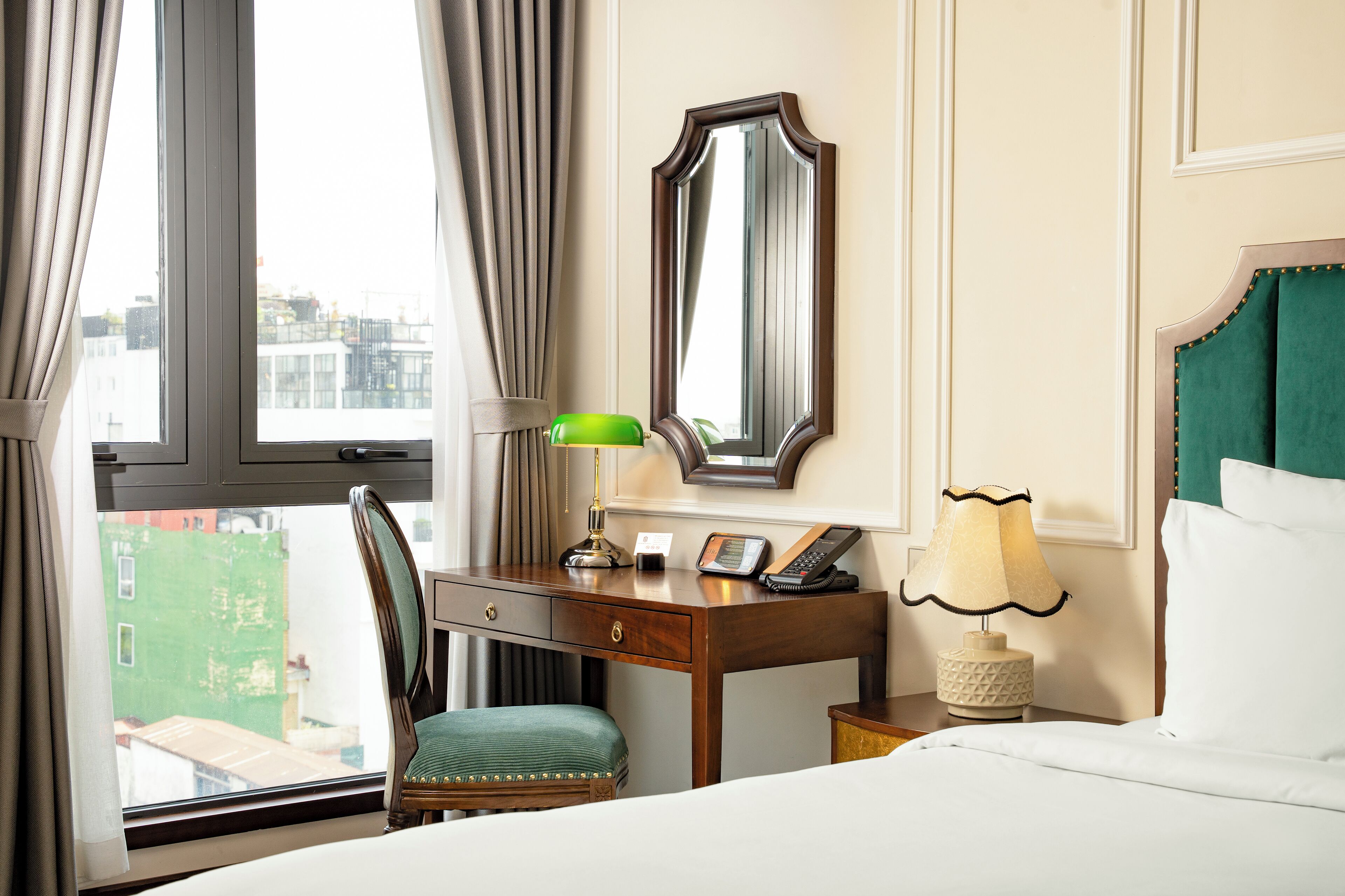 A hotel room with a bed, a desk with a telephone, a lamp, a chair, and a large mirror on the wall.