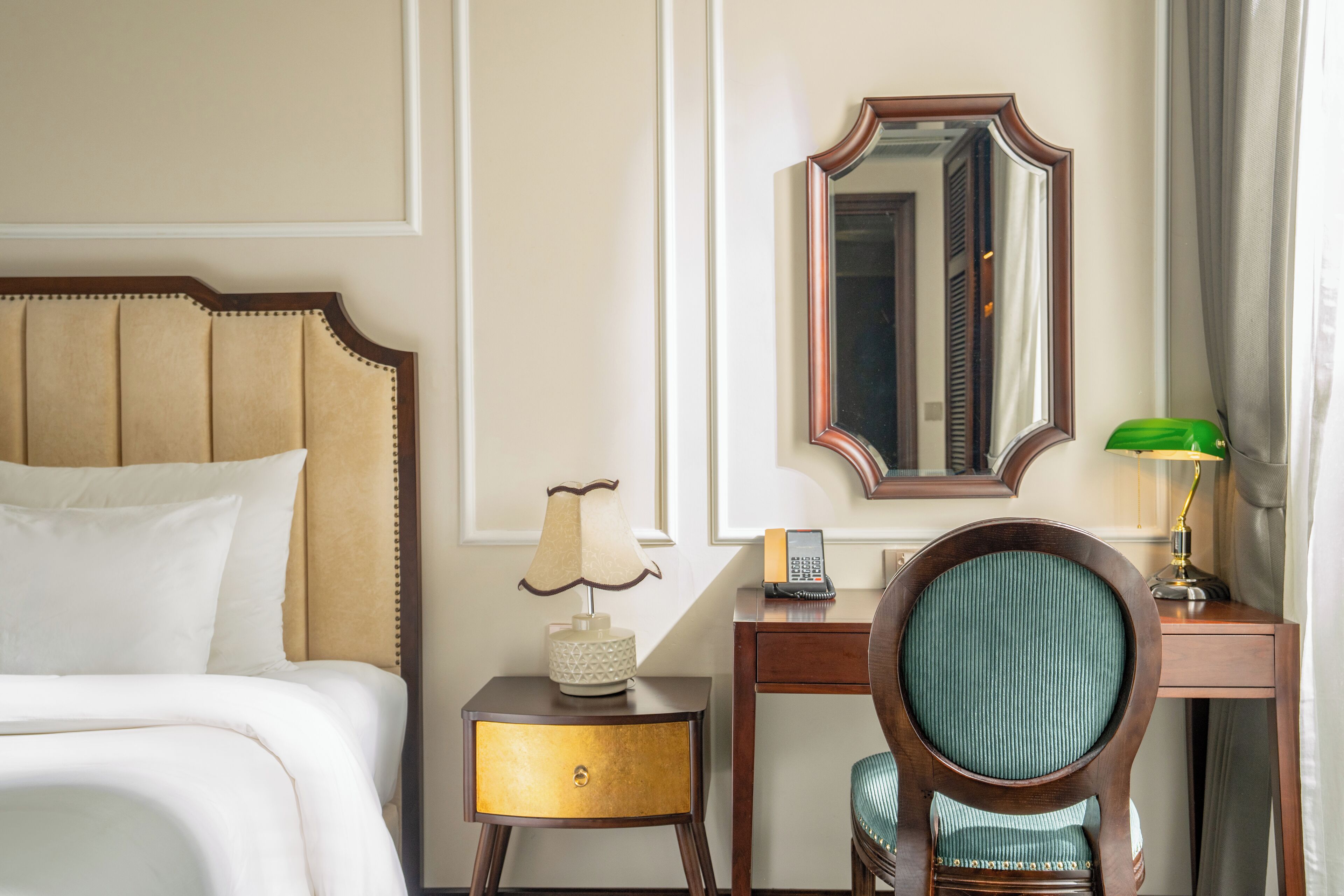 A hotel room with a bed, a nightstand with a lamp, a desk with a chair, and a mirror on the wall.