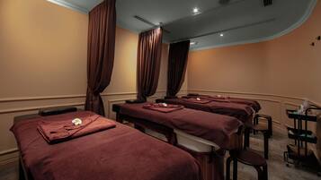 A room with three massage tables, each with a towel and a flower. There are curtains on the windows, and a cart with bottles and a container on the right.