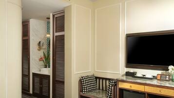 A hotel room with a television, a desk with a computer, a seating area with a patterned cushion, and a bathroom with a mirror and a plant.