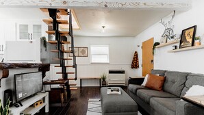 A modern living room with a grey sofa, a wooden staircase, and a kitchen area. - Cozy Lakeside Cabin with Hot Tub (Fawnskin)