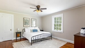 A bedroom with a bed, a ceiling fan, a window with blinds, and framed artwork on the wall.
