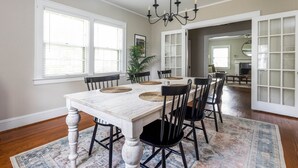 A dining area with a white table, black chairs, and a chandelier. There are large windows and a view into another room.