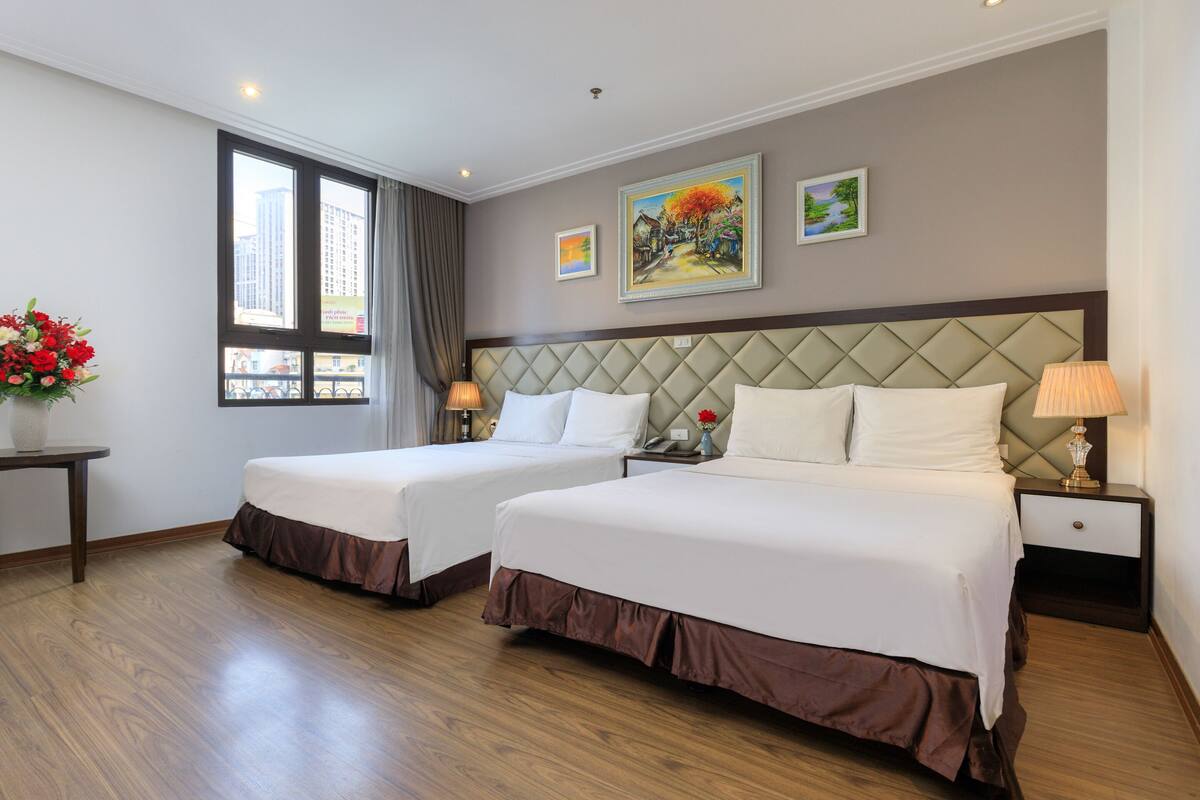 Habitación de hotel con dos camas, una ventana con vistas a edificios, un cuadro en la pared y una mesita de noche con una lámpara.