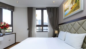 Une chambre d’hôtel avec un grand lit, une fenêtre avec des rideaux, un tableau au mur et un bureau agrémenté d’un vase de fleurs.