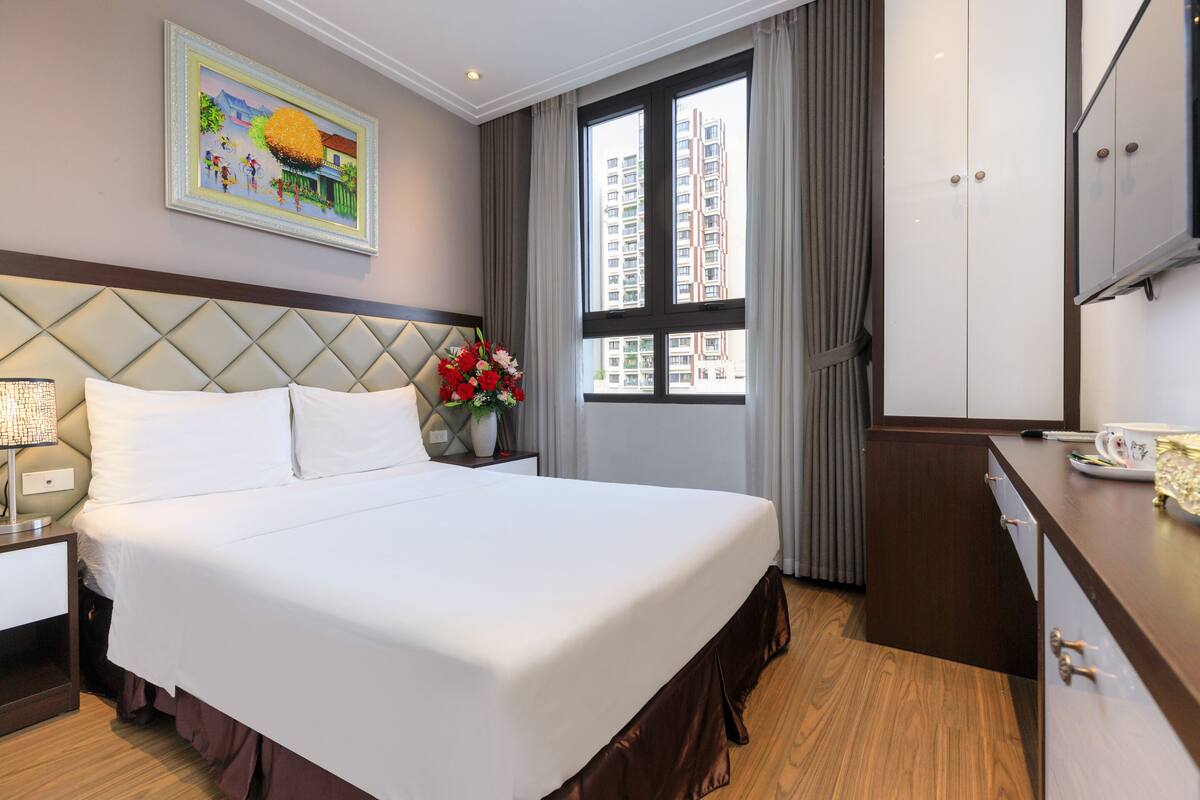 Una habitación de hotel con una cama grande, una mesita de noche con lámpara, un aparador, una ventana con cortinas y un cuadro en la pared.