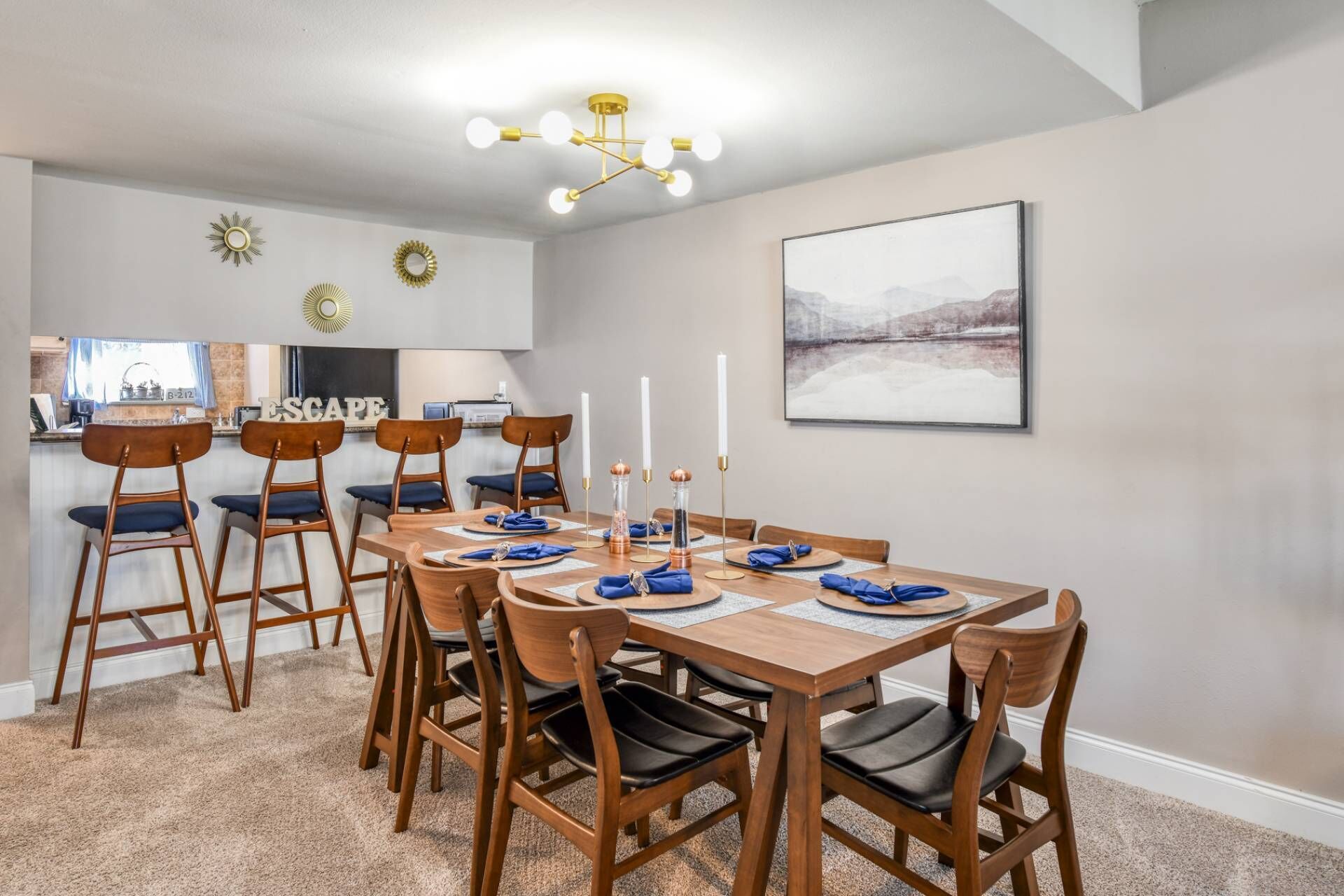 Un comedor con una mesa de madera dispuesta para seis personas, rodeada de sillas de madera con cojines azules. Un candelabro cuelga del techo y un cuadro con un paisaje está colgado en la pared.