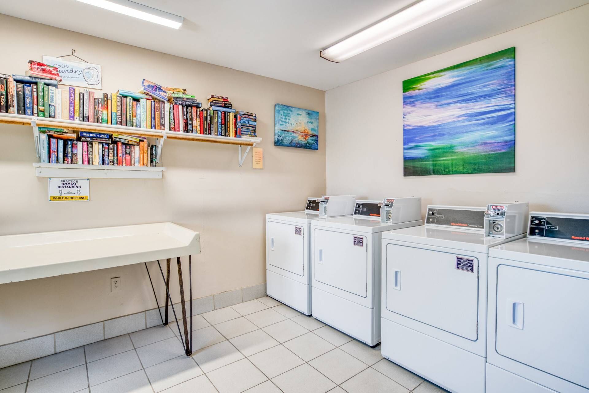 A laundry room with a white counter, multiple washing machines, a bookshelf with various books, and a painting on the wall.