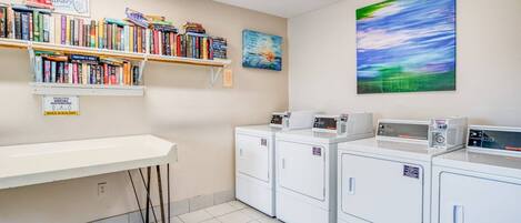 A laundry room with a white counter, multiple washing machines, a bookshelf with various books, and a painting on the wall.