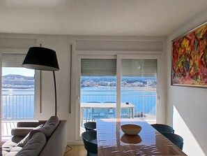 Ein modernes Zimmer mit einer Couch, einem Esstisch, Stühlen, einer Lampe und einem großen Fenster mit Blick auf ein Gewässer und Gebäude.