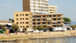 Un hotel de varios pisos con balcones, situado cerca de un cuerpo de agua con barcos y un litoral rocoso.
