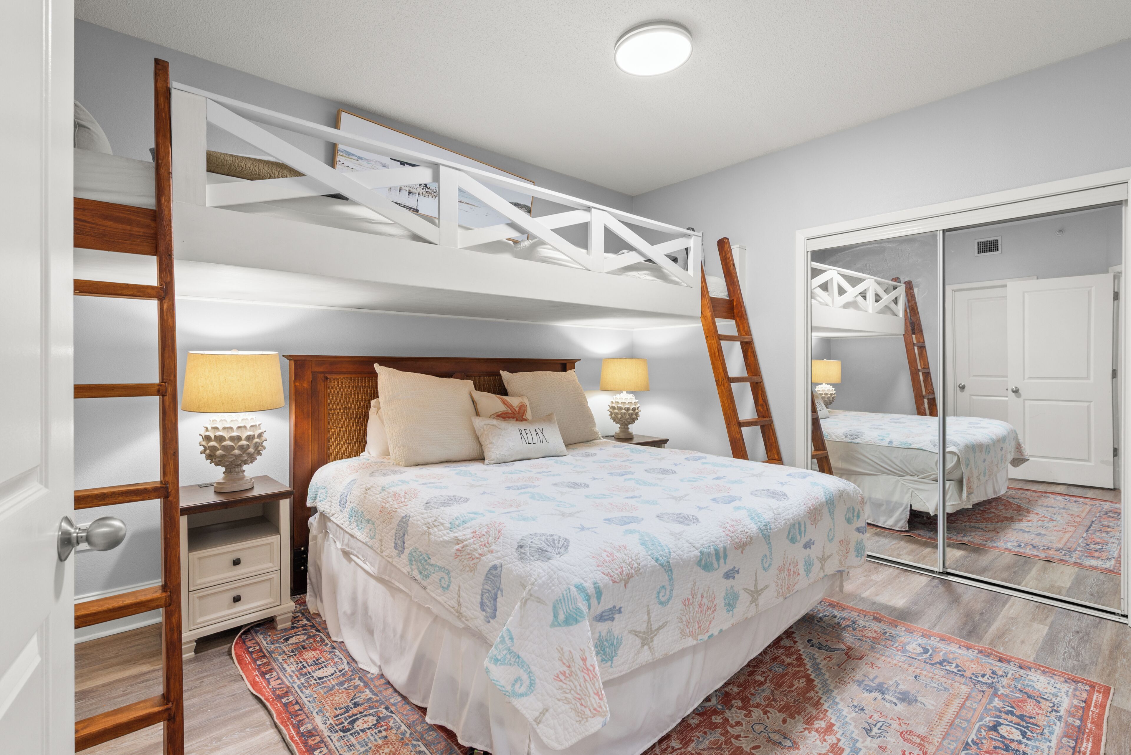 A bedroom with a bunk bed, a ladder, a bedside table with lamps, a mirror, and a patterned rug.