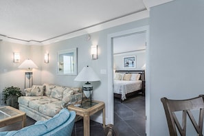 A living room with a sofa, a coffee table, and a lamp. There is a bed in the adjacent room.